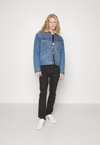 Blauwe spijkerjack met knoopsluiting, voorzien van borstzakken en een zigzagontwerp. G draagt over een grafisch zwart t-shirt en zwarte broek.