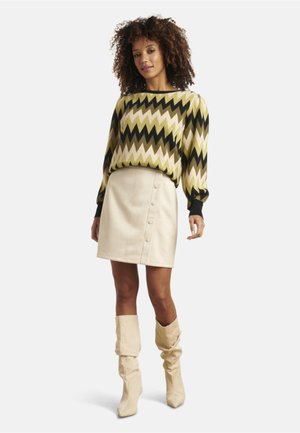 Pull avec un motif en zigzag vert, crème et noir, assorti à une jupe crème avec des boutons à l'avant. Bottes crème jusqu'aux genoux.