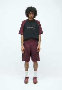 Schwarzes und kastanienbraunes Raglanarm-Shirt mit Timberland-Logo, kombiniert mit kastanienbraunen Cargo-Shorts, weißen Socken und kastanienbraunen Schuhen mit beigen Sohlen.