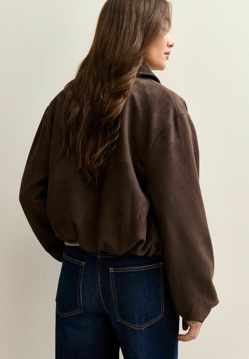 Chaqueta de ante marrón con un ajuste relajado, puños acanalados y un diseño corto, combinada con jeans oscuros. La vista posterior resalta la textura y la forma.