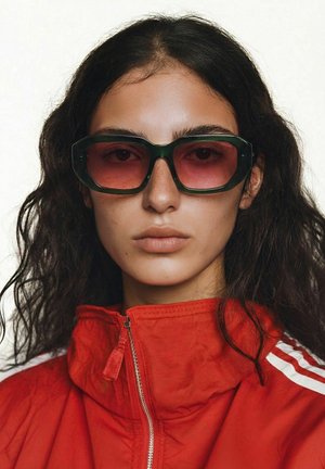 Femme aux longs cheveux bouclés foncés portant des lunettes de soleil surdimensionnées vertes et roses ainsi qu'une veste rouge zippée jusqu'au cou, sur un fond uni.