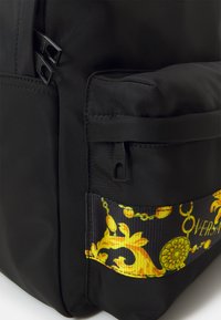 Sac à dos en tissu noir avec une finition brillante. Il présente une sangle décorative jaune et noire, ornée d'un motif en chaîne et de la marque Versace.