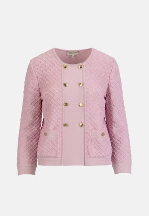 Žena má na sebe ružový sako s textúrou a zlatými gombíkmi, zodpovedajúce ružové nohavice, zlaté topánky a drží béžovú tkanú kabelku.