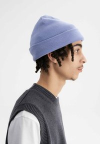 Lichtpaarse gebreide beanie met een omgevouwen rand en ribstructuur, gedragen door een persoon in een grijze mouwloze trui over een wit t-shirt.