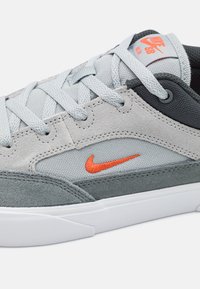 Sapatilha cinza em camurça e têxtil com o logótipo laranja da Nike, apresentando fecho com atacadores, lingueta almofadada e sola de borracha texturizada.