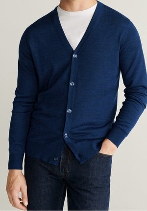 Uomo che indossa un cardigan blu navy abbottonato sopra una t-shirt bianca e jeans blu scuro, con una mano in tasca.