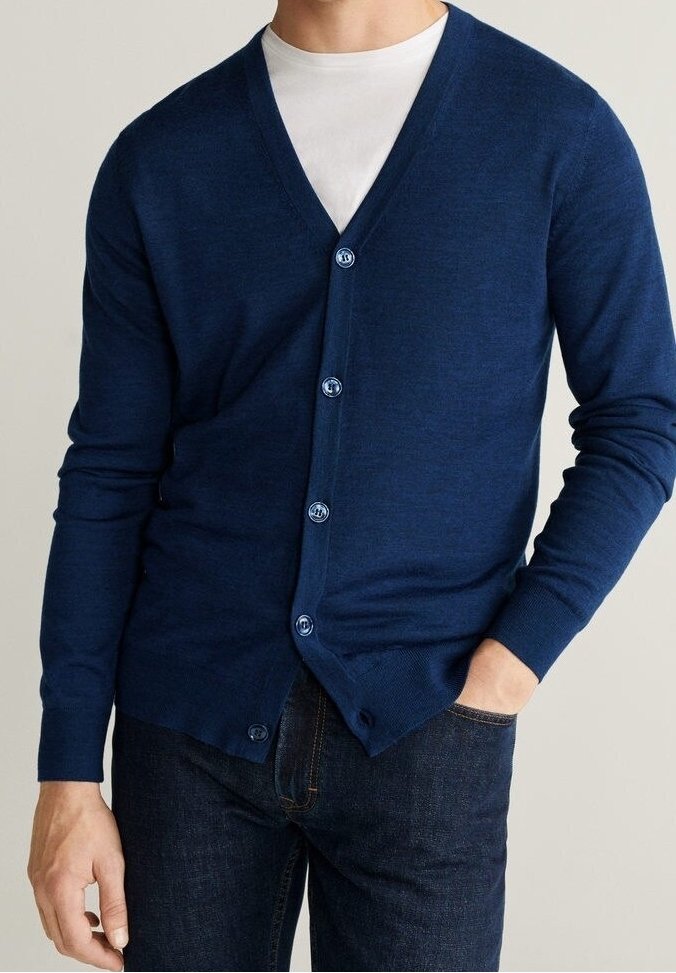 Homme portant un cardigan bleu marine boutonné par-dessus un t-shirt blanc et un jean bleu foncé, avec une main dans la poche.