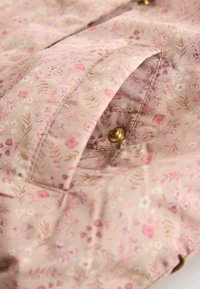 Textilie s květinovým vzorem na růžovém podkladu, která obsahuje malé květiny a listy; zahrnuje detail smosťovacího zlatého knoflíku.