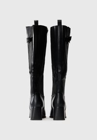 Par de botas de cuero negras hasta la rodilla con tacones de bloque y detalles de hebilla lateral, vistas desde la parte de atrás sobre un fondo liso.