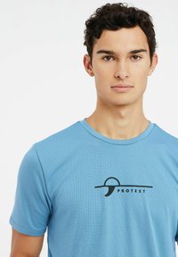 Mėlynas trumpomis rankovėmis sportinis marškinėliai, pagaminti iš tekstūruotos medžiagos, su juodu logotipu centre ir apvaliu iškirpimu.