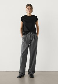 Žena stojící s rukama v kapsách, oblečená v černém krátkém tričku, šedých širokých kalhotách, černém opasku a černých botách na pozadí bílé zdi.