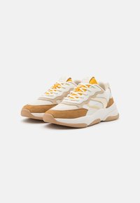 Pár béžových a bílých tenisek s akcenty z tan semiše, žlutým jazykem a bílými tkaničkami na bílém pozadí, nakloněné doprava.