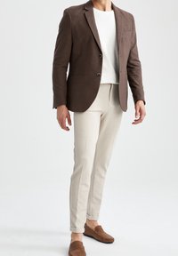 Uomo che indossa un blazer marrone, una camicia bianca, pantaloni beige slim-fit e mocassini marroni, in piedi davanti a uno sfondo bianco semplice.