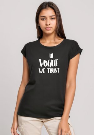 Young woman with long brown hair wearing a black t-shirt with white text "IN VOGUE WE TRUST" and beige pants against a plain background.