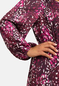 Fialové a růžové šaty se vzorem a elastickými balónovými rukávy. Hladký materiál s sklady a kontrastními bílými akcenty.