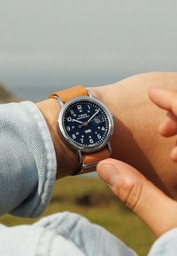Montre Timex Weekender avec un boîtier en acier inoxydable, un cadran bleu marine, des index en blanc et un bracelet en cuir marron. L'utilisateur règle l'heure.