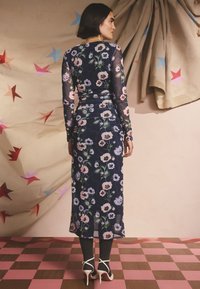 Ilga tamsiai mėlyna gėlėta suknelė su ilgomis rankovėmis, pagaminta iš permatomo audinio su rožinėmis, baltomis ir violetinėmis gėlėmis. Sijonas krenta žemiau kelių, derinamas su pilkomis kojinėmis ir baltomis aukštakulnėmis.