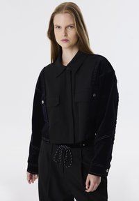 Zwarte, cropped jas met duale textuurstof; beschikt over twee borstzakken, knoopsluiting en contrasterende stiksels op de mouwen.