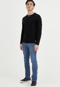 Schwarzes langärmliges Shirt mit V-Ausschnitt, kombiniert mit hellblauen Jeans und schwarzen Sneakers, vor einem weißen Hintergrund stehend.