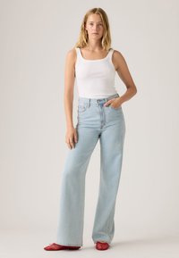 Γυναίκα που στέκεται με το χέρι στην τσέπη, φοράει λευκή φανέλα, ανοιχτό μπλε τζιν παντελόνι με φαρδιά πόδια και κόκκινα δίχτυα χαμηλά παπούτσια σε μονόχρωμο φόντο.