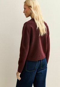 Burgundowa swetrowa bluzka o skróconym kroju, z miękką teksturą i subtelnymi detalami na ramionach, zestawiona z ciemnoniebieskimi dżinsami.