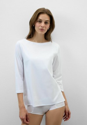 Frau mit langen braunen Haaren trägt ein weißes Langarmshirt und weiße Shorts und steht vor einer schlichten hellgrauen Wand.