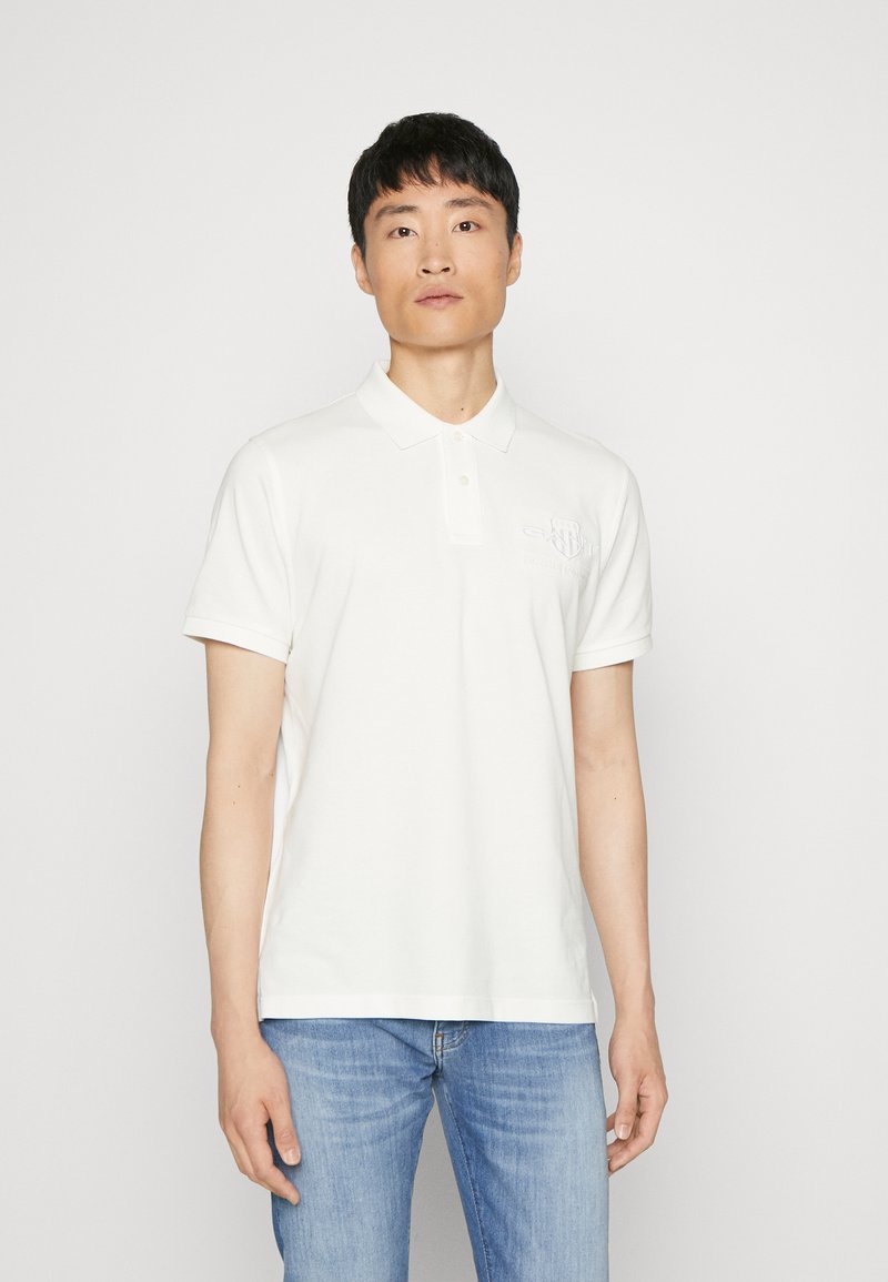 Polo shirt blanc avec un col et des manches courtes, fabriqué en coton. Dispose d'un logo brodé discret sur la poitrine gauche.