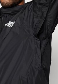 Giacca leggera nera con tasca con zip e un design minimalista. Il logo "The North Face" è visibile nella zona del petto.