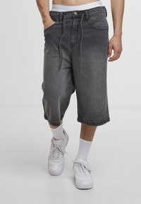 Άνδρας που φορά χαλαρά, ξεθωριασμένα μαύρα denim σορτς μέχρι το γόνατο με κορδόνι στη μέση, λευκές κάλτσες και λευκά αθλητικά παπούτσια, όρθιος με το ένα χέρι στην τσέπη.