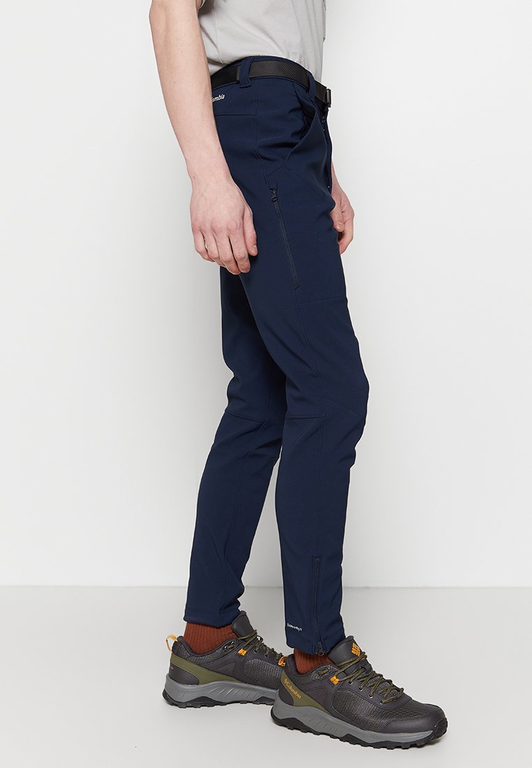 Pantalones azul marino, resistentes al agua, con un corte slim, que cuentan con un bolsillo lateral con cremallera y parches reforzados en las rodillas, combinados con zapatos de senderismo grises.