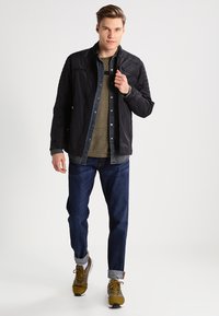 Chaqueta ligera negra llevada sobre una camisa verde y jeans azules. La chaqueta tiene un bolsillo con cremallera y una textura suave.