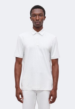 Homme portant un polo blanc à manches courtes avec un motif subtil et un pantalon blanc, debout devant un fond clair uni.