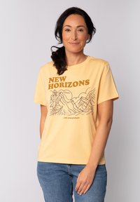 Geel katoenen T-shirt met korte mouwen, voorzien van bruine graphics van bergen en de tekst "NIEUWE HORIZONS" erboven en "...zijn overal" eronder.