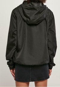 En person iklädd en svart huva-jacka och en mörk denimkjol, sedd bakifrån mot en enfärgad bakgrund.