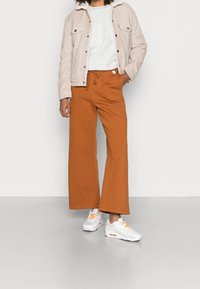 Brązowe szerokie dresy, jasnoszary t-shirt, beżowa kurtka z kordurowego materiału i białe sneakersy z pomarańczowymi akcentami. Gładka tekstura, luźny fason.