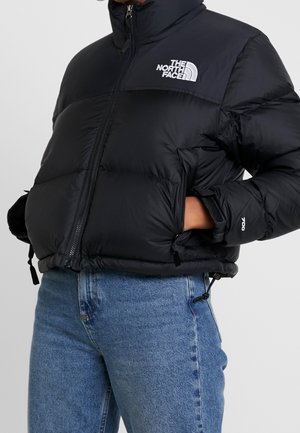 Czarna pikowana kurtka z wysokim kołnierzem, przednim zamkiem błyskawicznym i detalem z logo. Posiada boczne kieszenie oraz regulowany dół, zestawiona z niebieskimi dżinsami.