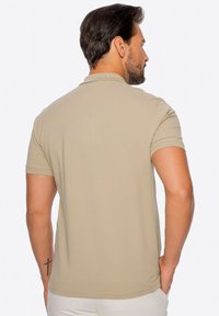 Béžové polo tričko s krátkým rukávem, ribbovanou texturou a plochým límcem. Obsahuje malé logo na horní části zad. Vhodné pro běžné nošení.