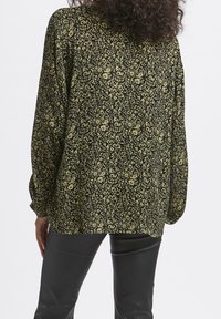 Blouse ample noire avec un motif floral vert et jaune. Manches longues avec des poignets élastiques, tissu fluide et ourlet arrondi.