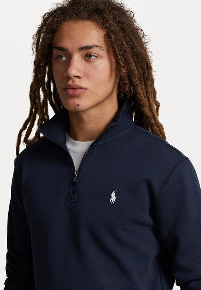 Marineblå zip-up hettegenser laget av mykt stoff, med høyt krage og hvit logo på venstre bryst. Teksturert overflate.
