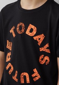 Zwarte katoenen T-shirt met een cirkelvormig ontwerp en gebarsten oranje tekst met de tekst "DE TOEKOMST VANDAAG." Ronde hals en korte mouwen.