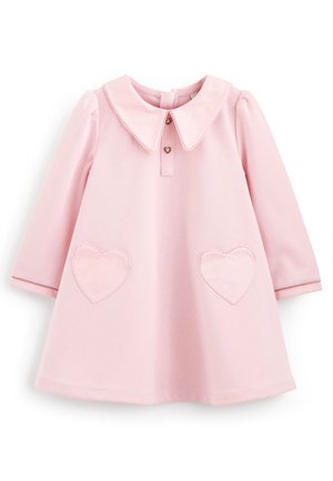 Niña pequeña caminando con un vestido rosa de manga larga con bolsillos en forma de corazón y zapatos blancos con grandes lazos rosas.