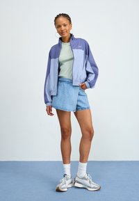 Giacca a vento lilla e blu, T-shirt verde, gonna di jeans, sneakers grigie con suole bianche, abbinate a calzini bianchi alla caviglia.