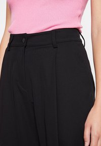 Pantalon noir taille haute avec une texture lisse, doté d'une fermeture à boutons sur le devant, de poches latérales et de détails délicats de plis.