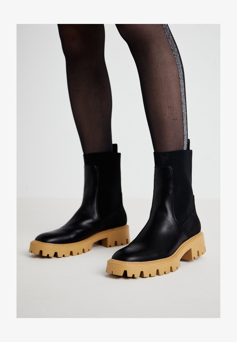 Botas de tornozelos em couro preto com sola de borracha bege robusta e detalhes em tecido canelado. Textura suave, bico arredondado e cano elástico alto.