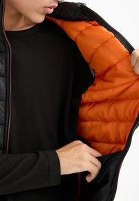 Chaqueta acolchada con un exterior negro y un forro naranja brillante. Las características incluyen un cierre de cremallera y un bolsillo con cremallera en el interior.