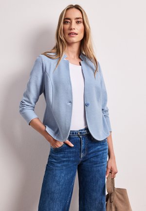 Frau mit langen blonden Haaren, die einen hellblauen strukturierten Blazer, ein weißes Hemd, blaue Jeans trägt und eine beige Wildlederhandtasche hält.