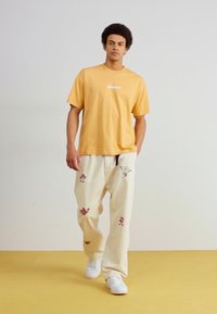 Tricou galben din bumbac cu logo alb; pantaloni crem cu broderie florală colorată; purtați cu adidași albi pe un podea galbenă.