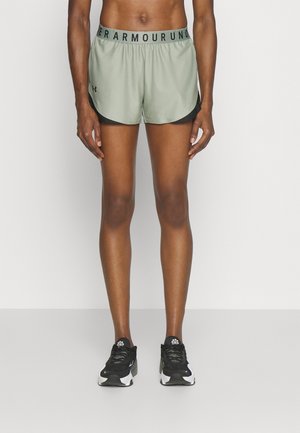 Slank person iført lysegrønne Under Armour løbeshorts og sort-hvide atletiksko står mod en ensfarvet baggrund.