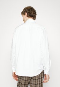 Chemise blanche à manches longues vue de dos, avec un col boutonné, un tissu lisse et un ourlet arrondi, associée à un pantalon à carreaux.
