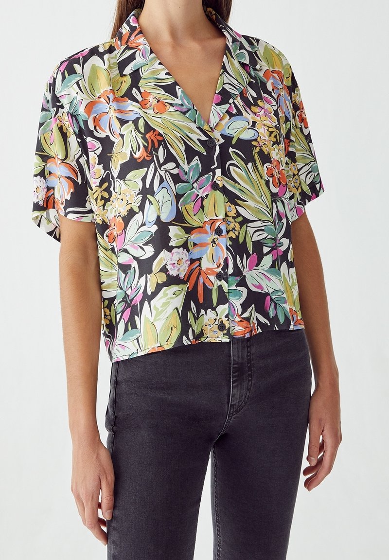 Femme portant une chemise noire à manches courtes avec imprimé floral coloré et un jean noir sur un fond clair uni.